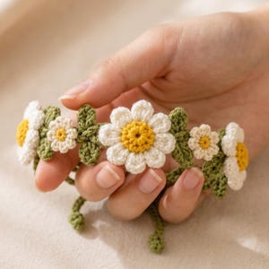 May include: A handmade crochet bracelet featuring daisy flowers. The bracelet has white petals with yellow centers, green leaves, and a green stem. The bracelet is held in a hand.