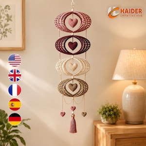 Puede incluir: Un móvil colgante vertical con cuatro anillos ovalados de ganchillo apilados en tonos rosa empolvado, burdeos oscuro, crema y bronceado. Cada anillo contiene un pequeño corazón de ganchillo a juego suspendido en el centro, con una borla.
