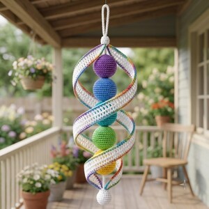 May include: A hanging crochet mobile featuring a central vertical column of five spheres in purple, blue, teal, green, and yellow. The spheres are encased in a white spiral ribbon with rainbow-colored edges, suspended by a white crochet loop.