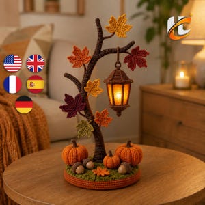 May include: A crocheted autumn-themed display featuring a dark brown tree trunk with orange, red, yellow, and green maple leaves. A hanging lantern with a warm yellow light is suspended from a branch. Two orange pumpkins and two acorns sit at the base.