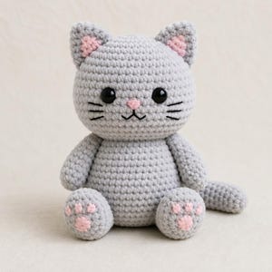 May include: A handmade grey crochet cat doll with pink detailing. The cat features black eyes, whiskers, and a pink nose. The ears and paw pads are pink. The cat is sitting up, displaying its detailed crochet texture.