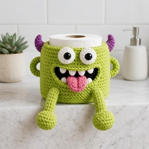 Peut inclure: Un porte-papier toilette monstre crocheté vert citron avec de grands yeux blancs, une bouche noire et une langue rose. Le support a des cornes, des bras et des jambes violettes. Un rouleau de papier toilette blanc est à l'intérieur.
