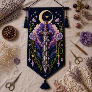 Könnte beinhalten: Ein marineblaues, kreuzgesticktes Wandbehang mit einem Schwertdesign, Mond und Sternen. Das Kunstwerk weist lila, lavendelfarbene und goldene florale Akzente auf, mit einer Quaste unten. Getrocknete Blumen und Bastelzubehör sind ebenfalls im Bild zu sehen.