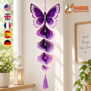 Könnte beinhalten: Ein hängendes Häkel-Mobile mit einem großen violetten Schmetterling an der Spitze, der mit weißen Perlen verziert ist. Unter dem Schmetterling hängt eine Reihe spiralförmiger, violett-ombréfarbener Häkelelemente an einer zentralen Perlenschnur mit Quaste.