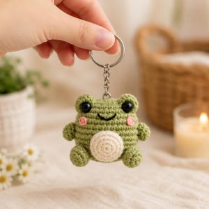 May include: A handmade green crochet frog keychain with black eyes and pink cheeks. The frog has a white belly and is attached to a silver keyring. The frog is held up by a hand.