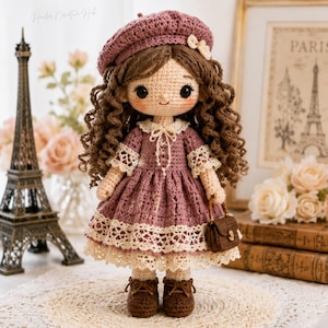 May include: A handcrafted crochet doll with brown curly hair, dressed in a mauve dress, beret, and shoes. The doll carries a small purse and features cream-coloured lace trim. The background includes a miniature Eiffel Tower and a framed Paris print.
