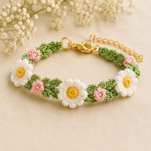 Puede incluir: Una pulsera de ganchillo hecha a mano con margaritas blancas con centros amarillos y pequeñas flores rosas, sobre hojas verdes. La pulsera tiene un cierre y extensor de cadena dorados. El diseño floral está sobre una base verde.