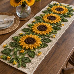 May include: A rectangular crochet table runner featuring a cream-colored base with a central row of three-dimensional sunflowers. The sunflowers have dark brown centers and bright yellow petals, surrounded by textured green leaves and small buds.