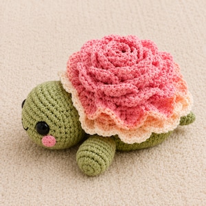 Könnte beinhalten: Eine handgefertigte gehäkelte Schildkröte mit einem großen, rosafarbenen rosenförmigen Panzer. Der Körper der Schildkröte ist grün, mit schwarzen Augen und einer rosa Wange. Der Rosenpanzer hat Schichten aus Rosa und Pfirsich.
