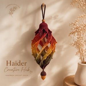 May include: A hanging crochet ornament featuring a spiral, cage-like structure with a gradient of colours transitioning from deep burgundy at the top to orange, yellow, and olive green at the bottom, finished with a small acorn-shaped tassel.
