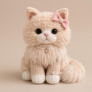 May include: A handmade crochet cat in beige with a pink bow, black eyes, and a white lace collar with a heart pendant. The cat has a fluffy texture and is wearing a silver ring with a pearl. The cat is sitting upright.