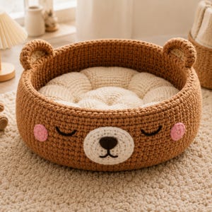 May include: A brown crochet pet bed shaped like a bear's face. The bed has a cream-colored cushion inside and pink cheeks. The ears are on the top of the bed. The bed is sitting on a beige carpet.