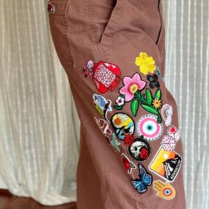 May include: Brown pants with multiple embroidered patches. Patches include flowers, a butterfly, a skull, and the word "Adventure". The pants have a wide leg and a classic five-pocket design.