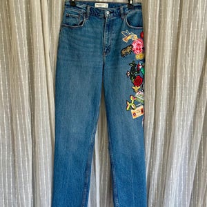 May include: Medium-wash blue denim jeans featuring a high-waisted fit, a button and zipper fly, and a raw-edge hem. The left leg is decorated with a vertical arrangement of colorful embroidered patches, including a bird, tiger, rose, and cassette.