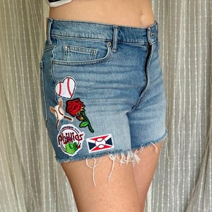 May include: Light blue denim shorts with frayed edges, featuring embroidered patches of a baseball, a heart, a rose, and a Phillies logo. The shorts have a classic five-pocket design.