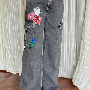 May include: Gray denim cargo pants with multiple pockets and decorative patches. Patches include a pink heart, a blue rose, and text that reads "FUNK IS DEAD" in red. The pants are wide-legged and worn with white sneakers.