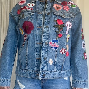 May include: A light-wash denim jacket adorned with colorful embroidered patches. Patches include floral designs, eye motifs, and the Phillies baseball team logo. The jacket has a classic button-up design and is paired with denim shorts.