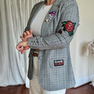 May include: A gray plaid blazer with patches and a red rose applique on the sleeve. The blazer has a "GAME OVER" patch on the pocket and decorative pins. The person is wearing a white t-shirt and striped pants.