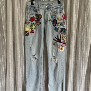 May include: Light blue denim jeans with multiple decorative patches. Patches include a red rose, a camera, a butterfly, and a cassette tape. The jeans have distressed details and slits at the ankles.