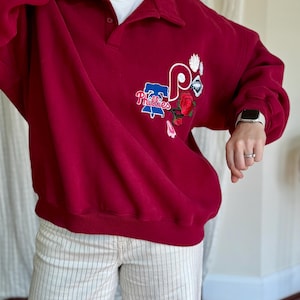 May include: A maroon sweatshirt with a collared neckline and the Philadelphia Phillies logo embroidered on the front. The logo includes the team name and a stylized "P" with floral accents. The sweatshirt is paired with striped pants.
