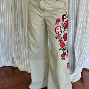 May include: Light green wide-leg jeans with a floral embroidery design on the left leg. The embroidery features red roses, green leaves, and a red and white patch with the letter "P".
