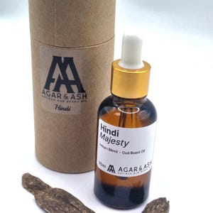 Puede incluir: Una botella de vidrio marrón de aceite para barba Hindi Majesty Artisan Oud con tapa dorada y cuentagotas blanco. Una caja cilíndrica de cartón y dos trozos de madera están junto a la botella. La etiqueta dice "AGAR & ASH".
