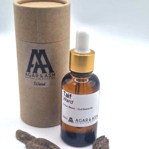 May include: A brown glass bottle of Taif Ward Artisan Blend Oud Beard Oil with a white dropper and gold cap. A cylindrical cardboard container and wood pieces are also present. The brand name is Agar & Ash.