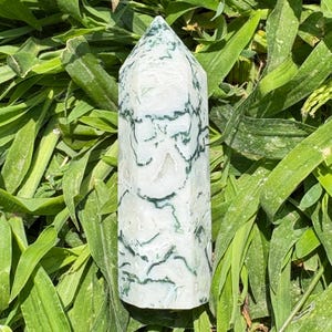 May include: A polished, six-sided crystal tower featuring a translucent white base with intricate, dark green moss-like inclusions throughout the stone. The mineral specimen has a pointed top and a smooth, cylindrical body with distinct natural patterns.