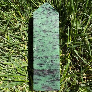 May include: A tall, rectangular green zoisite crystal tower with black speckles and streaks. The polished stone has a flat top and is set against a backdrop of green grass.