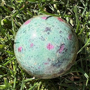 May include: A polished spherical stone featuring a mottled light green base color with scattered, distinct inclusions of vibrant magenta and dark black mineral deposits throughout the surface, creating a natural speckled appearance.