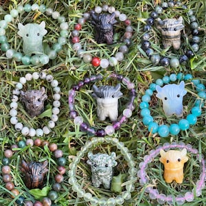 May include: A collection of beaded bracelets featuring carved stone bull figurines. The bracelets showcase a variety of colors, including green, black, white, and purple. The beads are made of various stones and are arranged in a circular pattern.