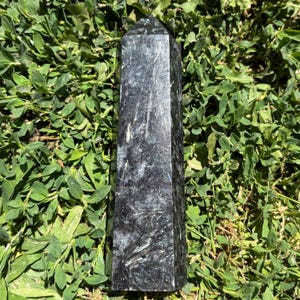 May include: A polished black and silver crystal tower with a hexagonal base and a pointed top. The stone features a dark, charcoal-colored surface with intricate, needle-like silver inclusions that create a shimmering, fibrous texture throughout.