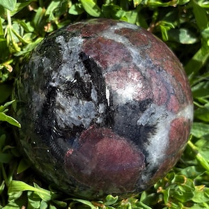 May include: A polished spherical stone featuring a mottled pattern of deep maroon, dark charcoal, and translucent white mineral inclusions. The surface is smooth and reflective, displaying a complex interplay of dark and light crystalline textures.