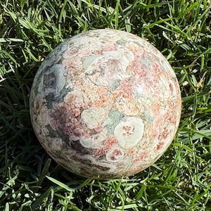 May include: A polished spherical stone featuring a complex, mottled pattern of earthy tones including shades of dusty rose, sage green, cream, and dark brown. The surface displays a natural, intricate texture with varied circular and irregular inclusions.