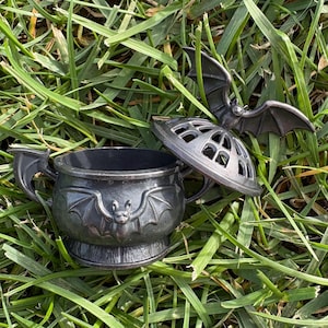 May include: A dark gray, metallic-toned cauldron-shaped container featuring embossed bat figures on the sides and handles. The matching lid is topped with a sculpted bat with outstretched wings and includes arched cutouts for ventilation.