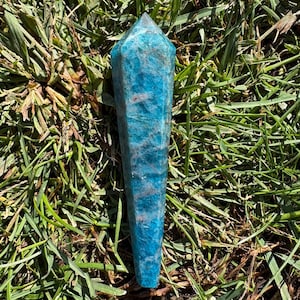 May include: A polished, hexagonal crystal point in a vibrant shade of blue-green. The stone features natural internal inclusions and a faceted, tapered top, showcasing a translucent quality with varied textures across its elongated surface.