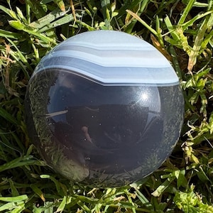 May include: A polished, spherical agate stone with bands of gray, white, and black. The gemstone is displayed on a bed of green grass, showcasing its natural colors and patterns. The smooth surface reflects light, highlighting the stone's texture.
