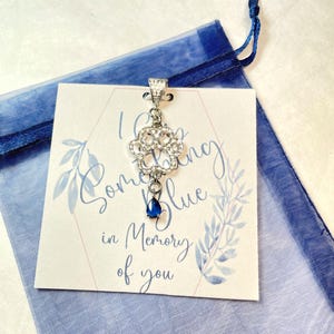 May include: A silver pendant with a blue teardrop-shaped stone hanging from a silver filigree design. The pendant is on a white card with the text "I Carry Something Blue in Memory of You".