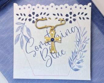 May include: A gold-colored cross pin with a blue gemstone center and white accents, attached to a safety pin. The pin is displayed on a white card with the words "Something Blue" and floral illustrations, set against a blue fabric background.