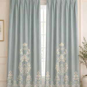Può includere: Una coppia di tende azzurre con un motivo damascato ricamato color crema lungo l'orlo inferiore che si estende verso l'alto. Le tende sono appese a un bastone in metallo dorato con anelli e presentano un design superiore plissettato.
