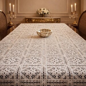 Può includere: Una tovaglia color crema, finemente lavorata all'uncinetto con un motivo floreale, copre un tavolo in legno scuro. Una ciotola d'argento è al centro. L'ambiente della sala da pranzo comprende due candelabri e un tavolo decorativo sullo sfondo.