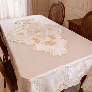 以下が含まれることがあります： クリーム色のテーブルクロス。金とクリーム色の花の刺繍が施され、木製のテーブルに掛けられています。テーブルクロスはスカラップエッジで縁取られ、ダイニングエリアに優雅さを添えています。テーブルの周りには木製の椅子が配置されています。