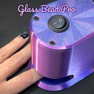 Puede incluir: Máquina Glass Bead Pro morada y azul con un interruptor de encendido/apagado negro. La máquina descansa sobre una superficie negra. El texto "Glass Bead Pro" está en la esquina superior izquierda.