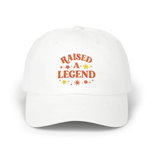 May include: White baseball cap with the embroidered text "RAISED A LEGEND" in orange and yellow. The text is surrounded by small flower and star designs. A casual hat for everyday wear.