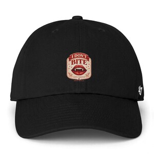 May include: Black baseball cap with a curved brim. The cap features a rectangular patch with the words "I DON'T BITE" above a graphic of red lips and fangs. The number "47" is embroidered on the side.