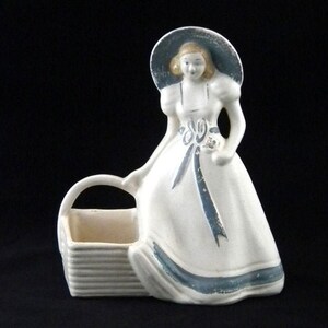 May include: A white ceramic figurine of a woman in a long dress and hat, holding a basket. The dress and hat have blue accents. The figurine is approximately 7 inches tall.