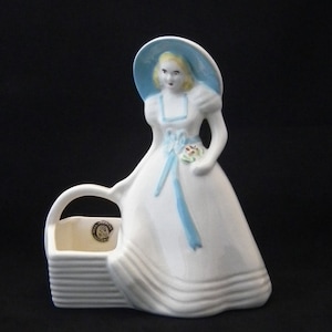 May include: A white ceramic figurine of a woman in a long dress and a blue hat. The figurine holds a small bouquet and has a basket attached. The figure has blonde hair and blue accents.
