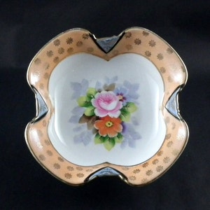 Puede incluir: Un pequeño plato decorativo de porcelana con un diseño floral. El plato tiene un interior blanco con un ramo de flores rosas, naranjas y moradas. El borde es de color melocotón con lunares dorados y un borde festoneado.