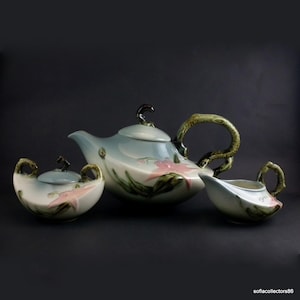 May include: A vintage ceramic tea set with a teapot, creamer, and sugar bowl. The set features a light blue and white color scheme with pink floral accents and green leaf details. The handles and finials have a textured, branch-like design.