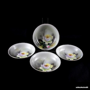 Puede incluir: Juego de cuatro cuencos de porcelana blanca. Cada cuenco presenta un diseño floral pintado a mano con una flor amarilla y morada. Los cuencos están dispuestos sobre un fondo negro, mostrando su elegante diseño.